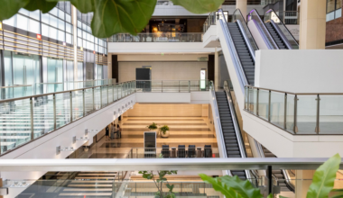 A modern, multi-level indoor space with escalators, glass railings, plants, and natural light from large windows.