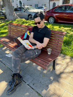A person sitting on a park bench reading a book, with a drink nearby.