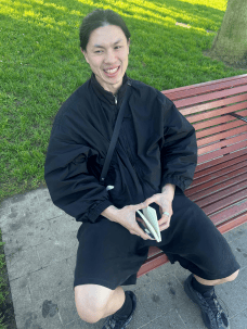 A person in black clothing sitting on a park bench, holding a small notebook.