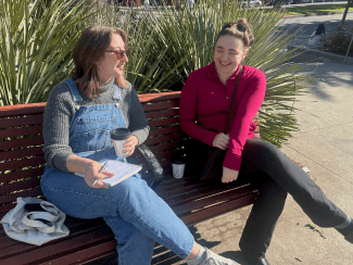 Two women sitting on a park bench talking and laughing while holding coffee cups.