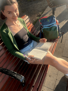 A young woman sits on a park bench, writing in an open notebook with a pen, with a backpack resting beside her.