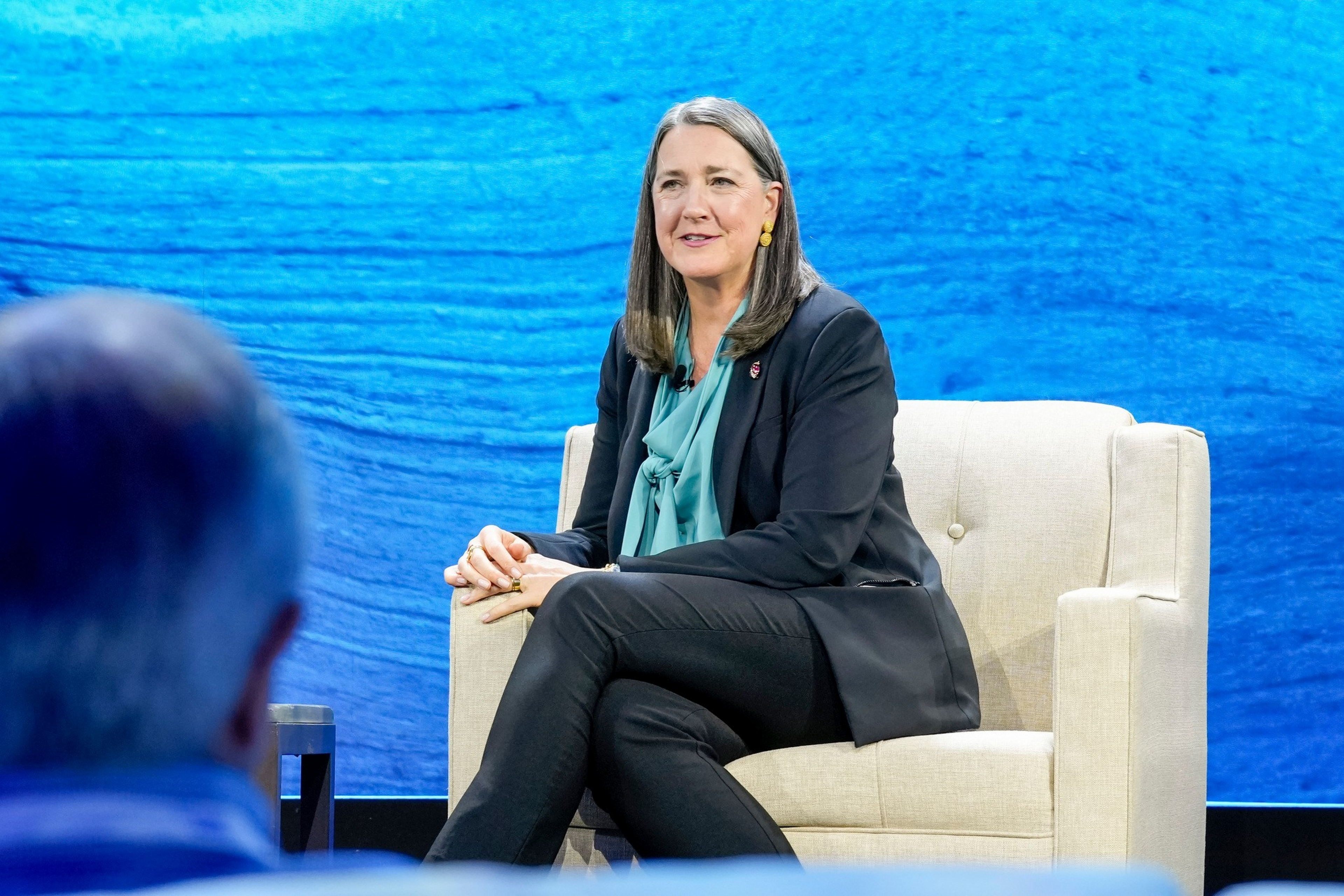 A woman in a black suit and teal blouse sits on a beige armchair against a blue textured backdrop, engaging in a conversation with a person in the foreground.