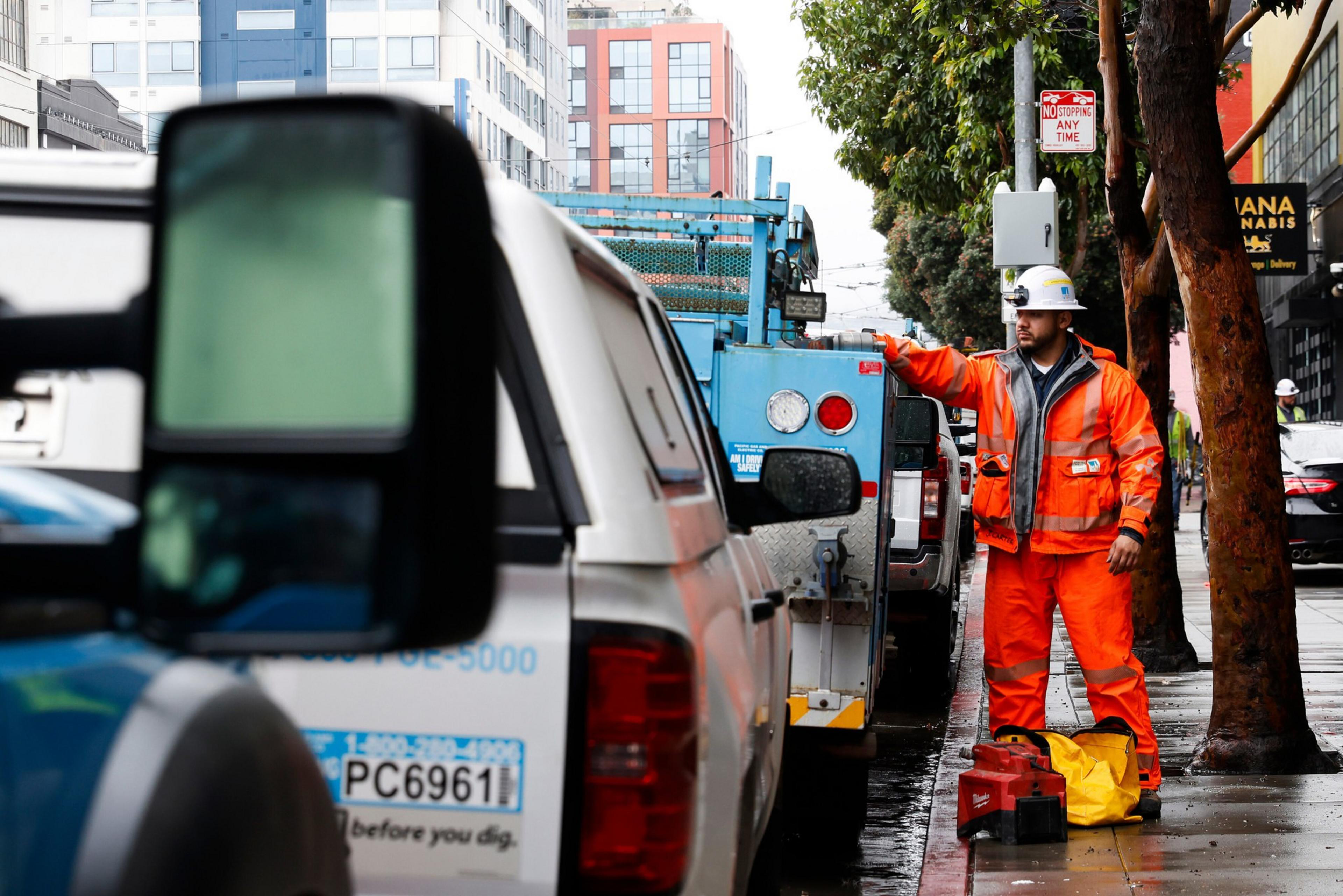 A worker in bright orange safety gear and a white hard hat stands on a wet sidewalk beside parked utility trucks on a city street.