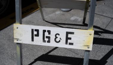 A wooden sign with black letters "PG&E" is attached to a metal frame on a concrete sidewalk.
