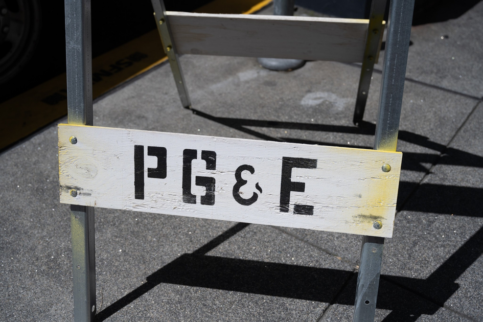 A wooden sign with black letters "PG&E" is attached to a metal frame on a concrete sidewalk.