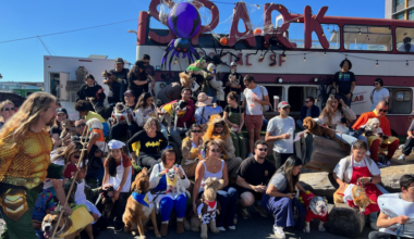 A large group of people and dogs, some in costumes, gather outdoors in front of a double-decker bus with Halloween decorations, including a large spider.