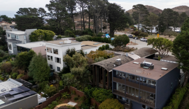 Residential houses surrounded by trees and greenery, with a road, hills, and tall pine trees in the background under a partly cloudy sky.