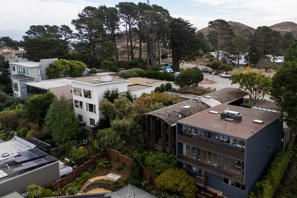 Residential houses surrounded by trees and greenery, with a road, hills, and tall pine trees in the background under a partly cloudy sky.