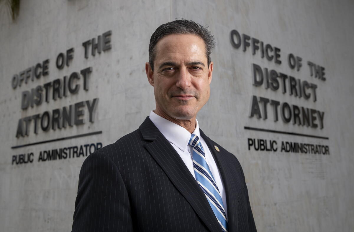 A man in a dark suit, with a blue striped tie, outside a building with signs that say Office of the District Attorney