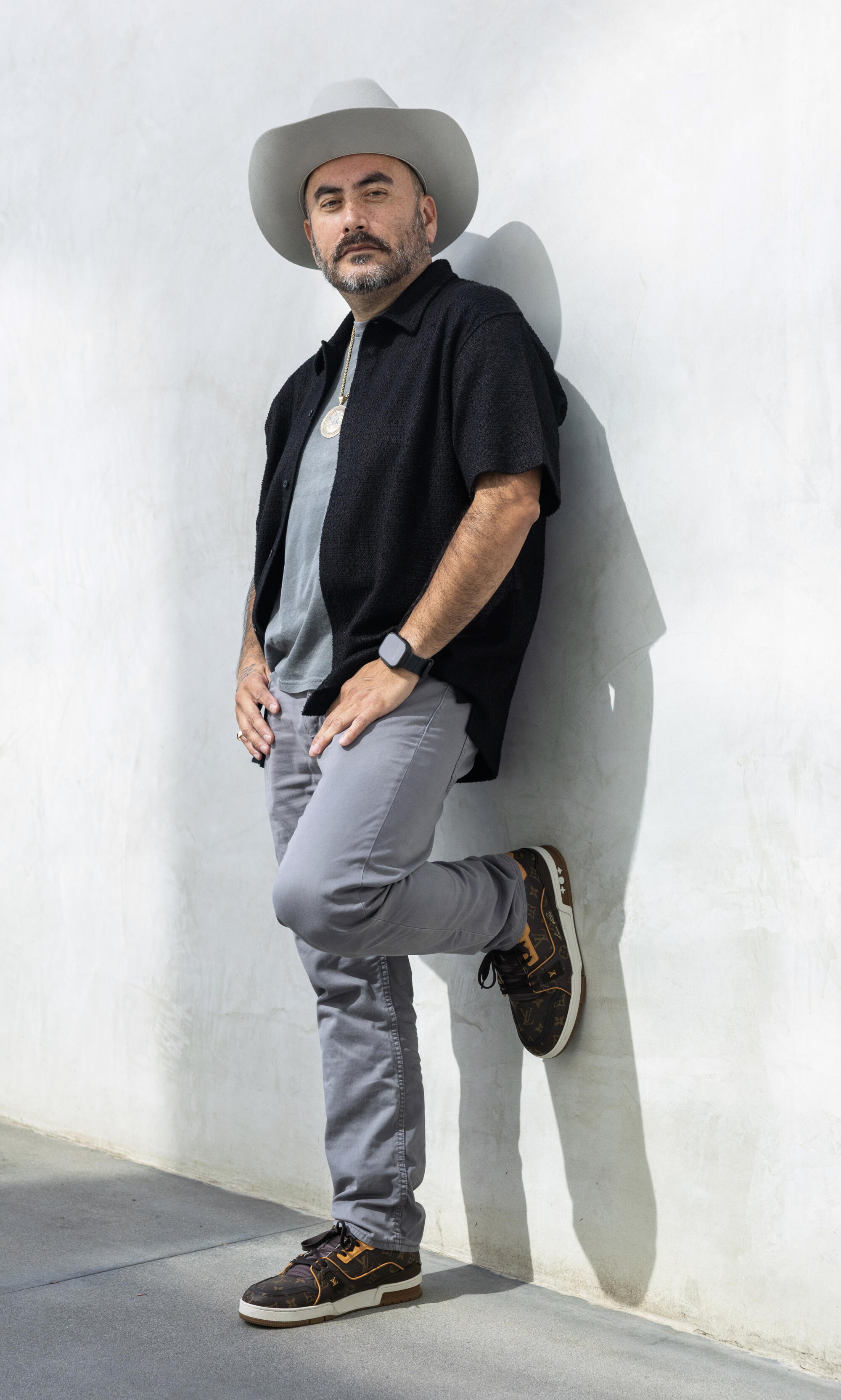 Comedian Alfred Robles stands against white wall with one foot up