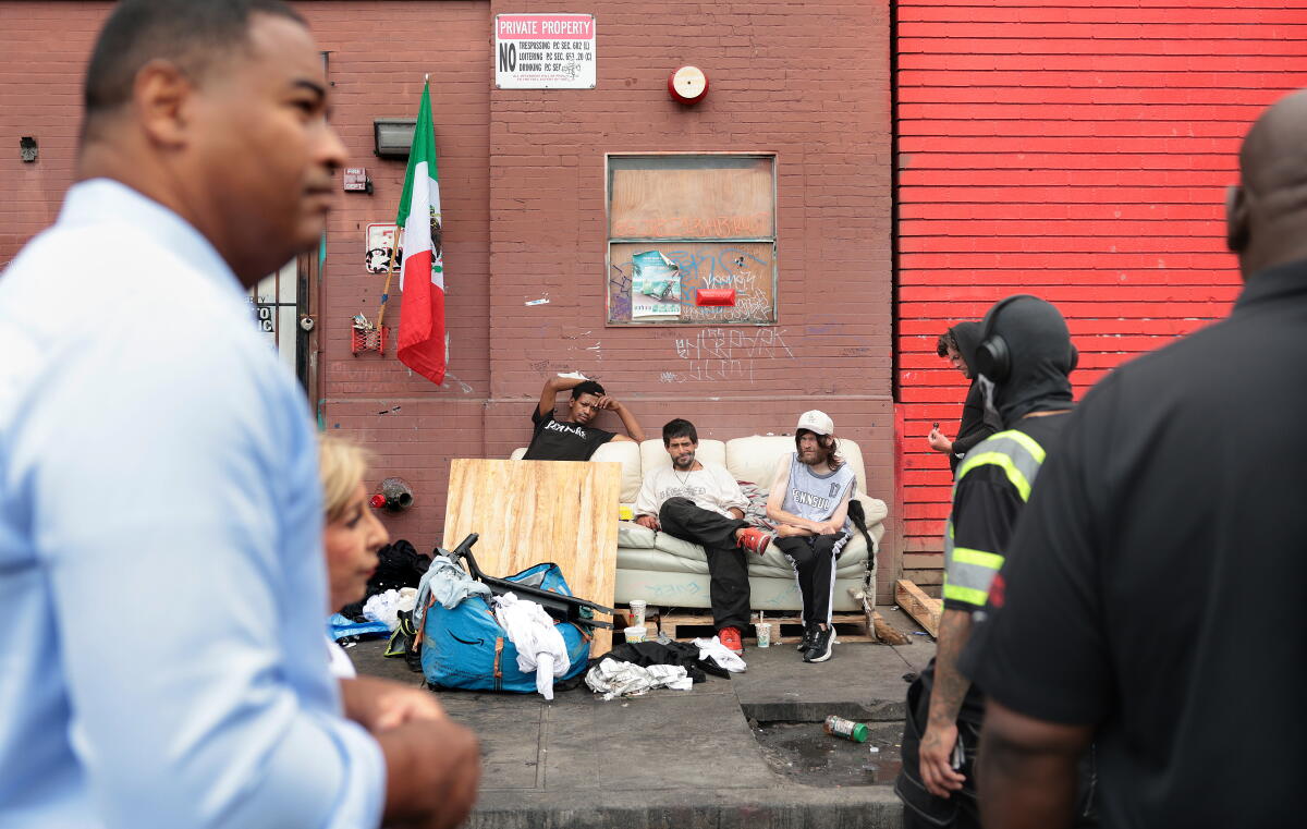 Three people sit on a couch on a sidewalk as several people watch from the street.