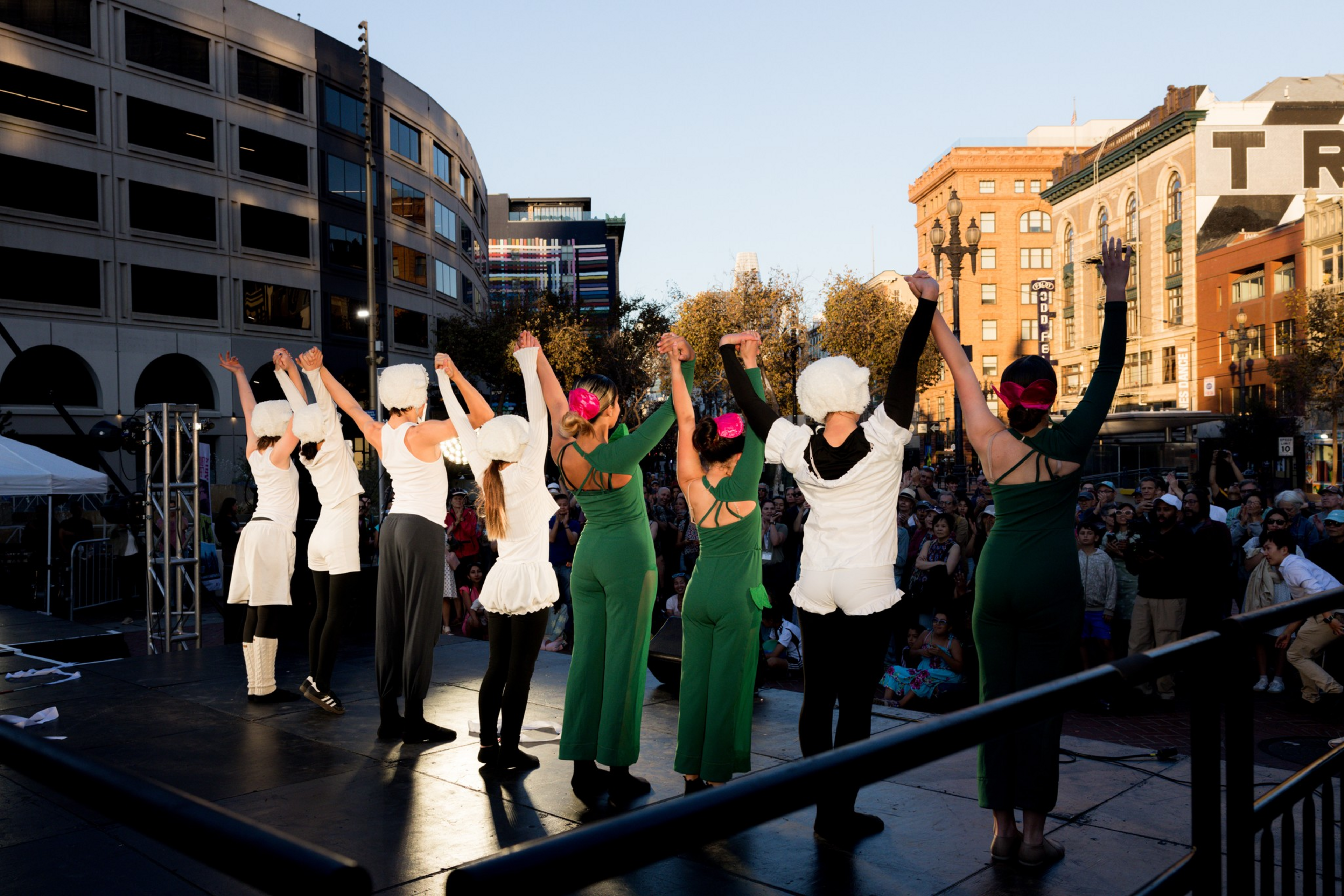 Eight performers stand on an outdoor stage facing a crowd, raising their hands in a bow; some wear white costumes and others green outfits.
