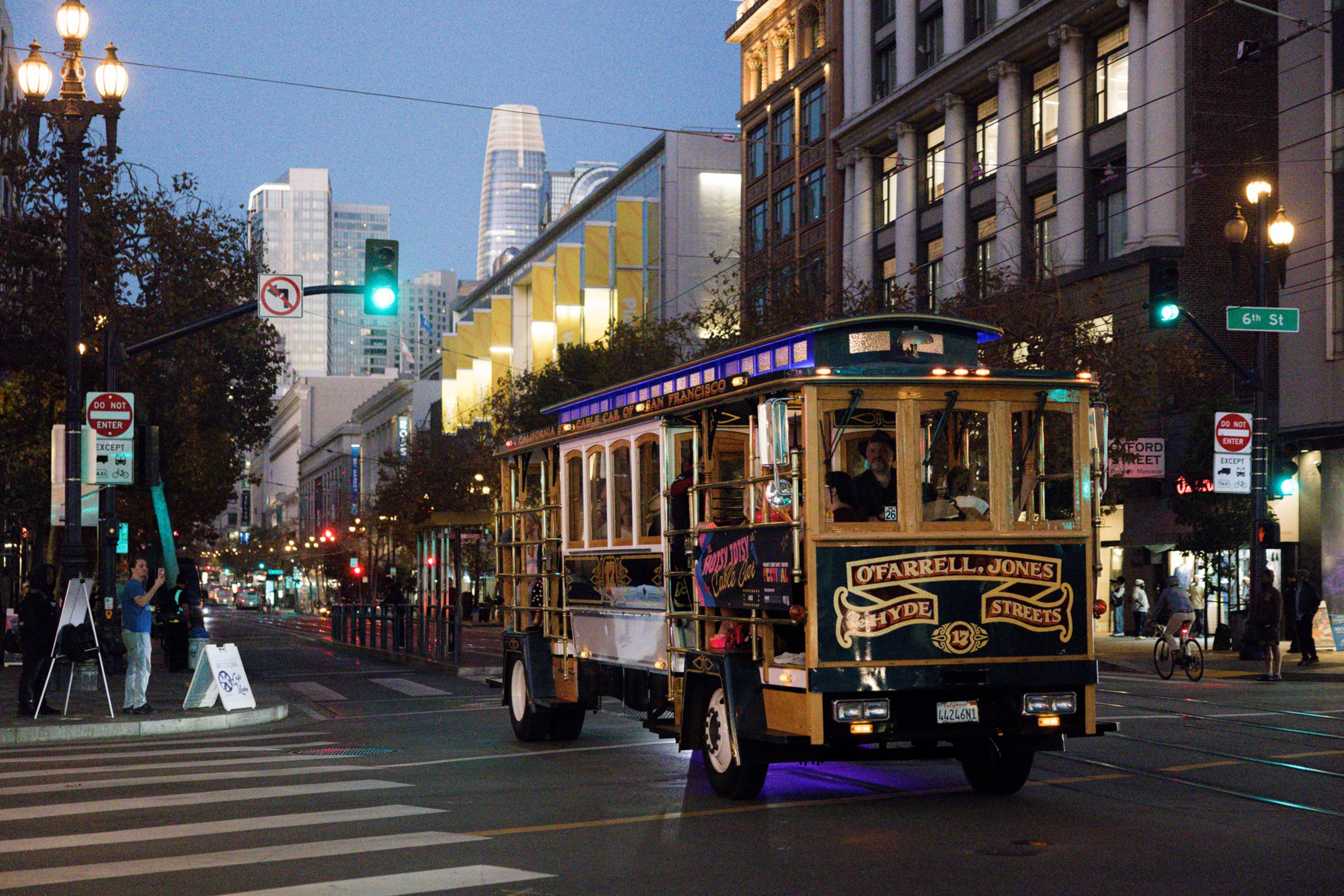 A San Francisco cable car with passengers travels on a city street at dusk, surrounded by tall buildings and illuminated street lamps.
