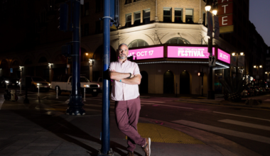 A man with a beard leans against a street pole at night near a theater with a bright marquee advertising a festival on October 17.