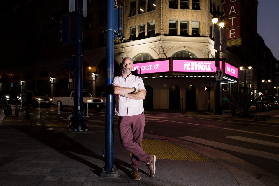 A man with a beard leans against a street pole at night near a theater with a bright marquee advertising a festival on October 17.