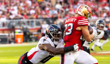 A football player in a red San Francisco 49ers uniform clutches the ball while a player in a white Atlanta Falcons uniform tackles him from behind.