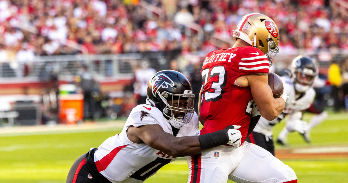 A football player in a red San Francisco 49ers uniform clutches the ball while a player in a white Atlanta Falcons uniform tackles him from behind.