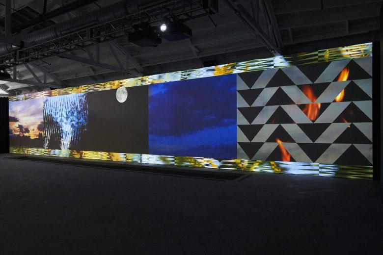 A large wall displays a digital triptych: an image of a dome structure under a sky, a dark night sky with a moon, and a geometric pattern with orange lights.