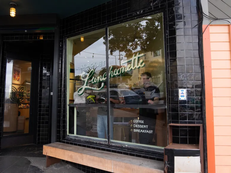The exterior of Black Jet Luncheonette with a bench and window decals reading “Coffee,” “Dessert,” and “Breakfast.”