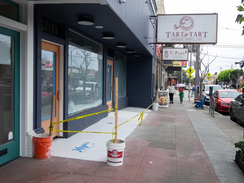 A storefront entrance under construction with caution tape, leading to a sidewalk with signs for Tart to Tart bakery and Yi Fang tea shop.