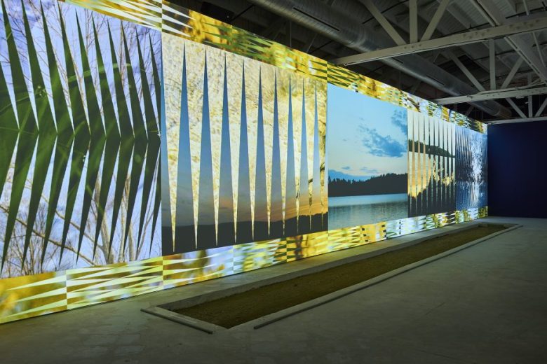 A large digital projection displays abstract and nature-themed images on a wall in a modern gallery with exposed ceiling beams and a rectangular recessed area in the floor.