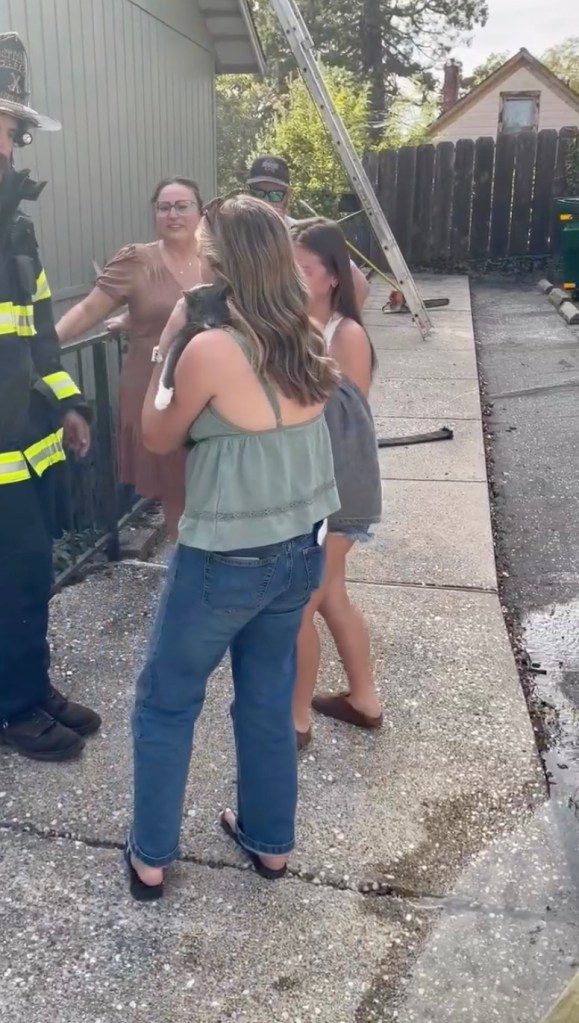 A woman holding a cat she was reunited with after it was rescued from a fire, standing with firefighters and family outside a home.