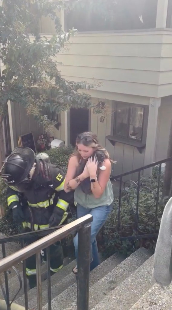 A firefighter in full gear and a woman holding a cat walk up exterior stairs outside an apartment building.