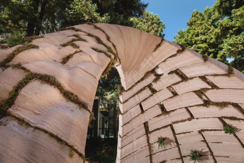 Curved structure made of layered, overlapping tiles embedded with moss, set outdoors among tall trees, with sunlight filtering through the foliage.