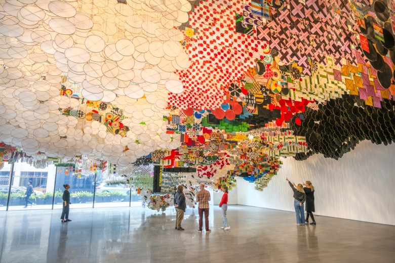 People stand under a large, colorful installation of hanging flags and circular shapes in an art gallery with floor-to-ceiling windows.