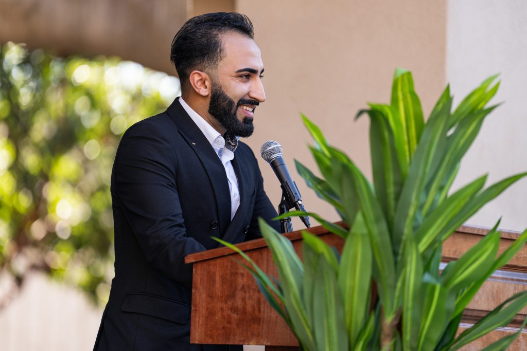 A person in a black suit stands at a wooden podium, speaking into a microphone with a smile, as green foliage frames the scene.