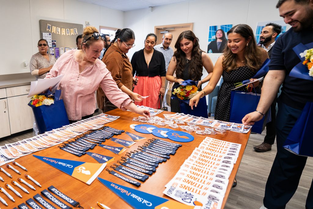 A group of people gather around a table filled with promotional items, including branded pens, flags, and gift bags, at what appears to be a welcome or orientation event.