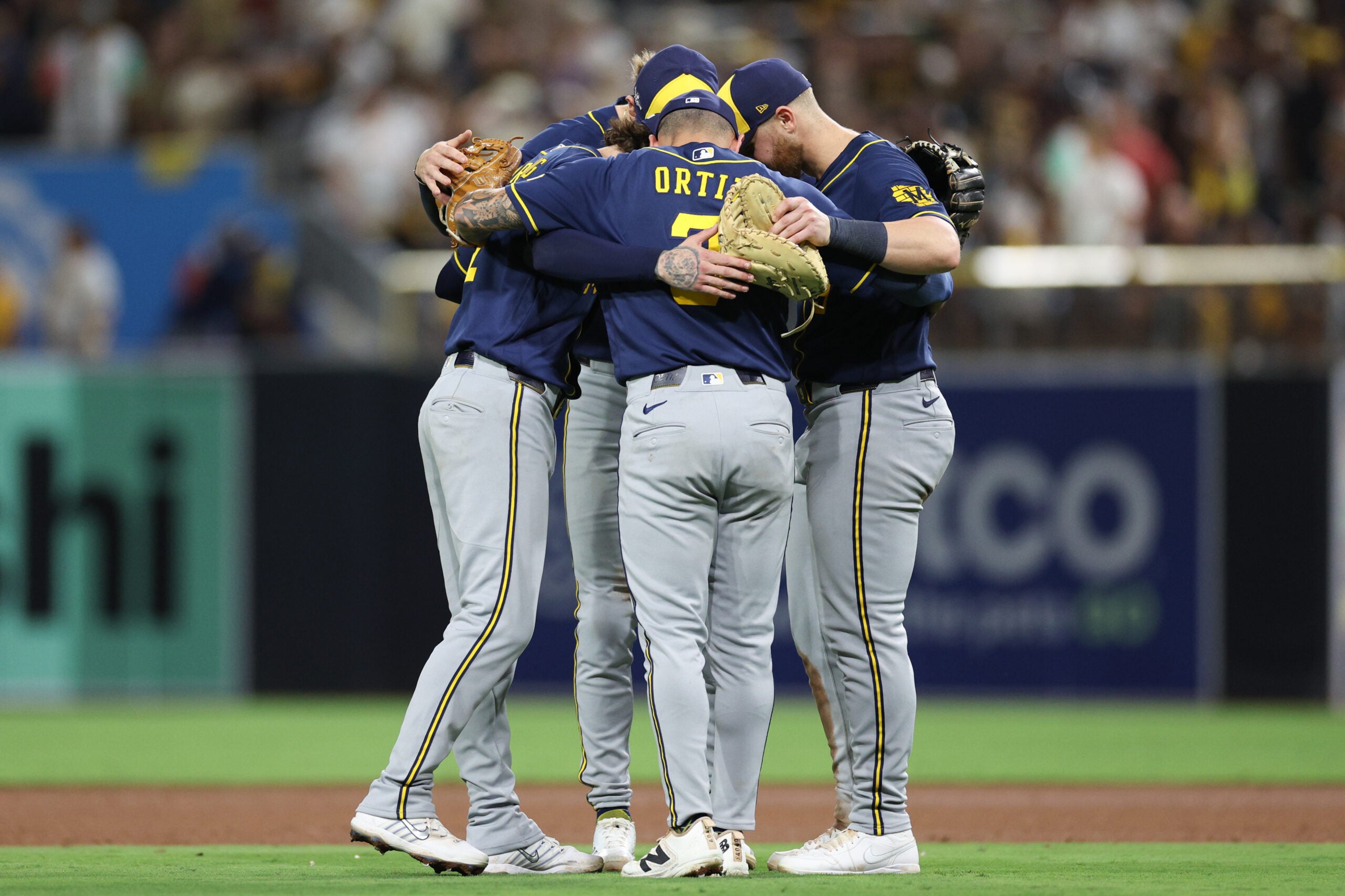 Four baseball players in Milwaukee Brewers uniforms huddle together on the field.
