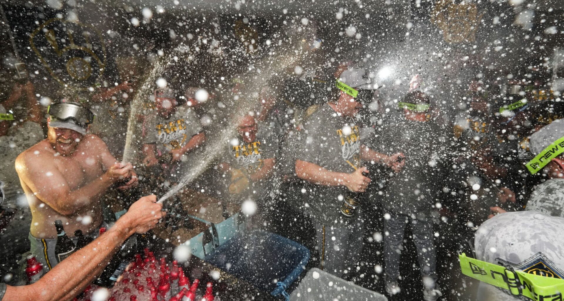 A group of people wearing matching gray shirts celebrate in a locker room as champagne is sprayed, filling the air with droplets.