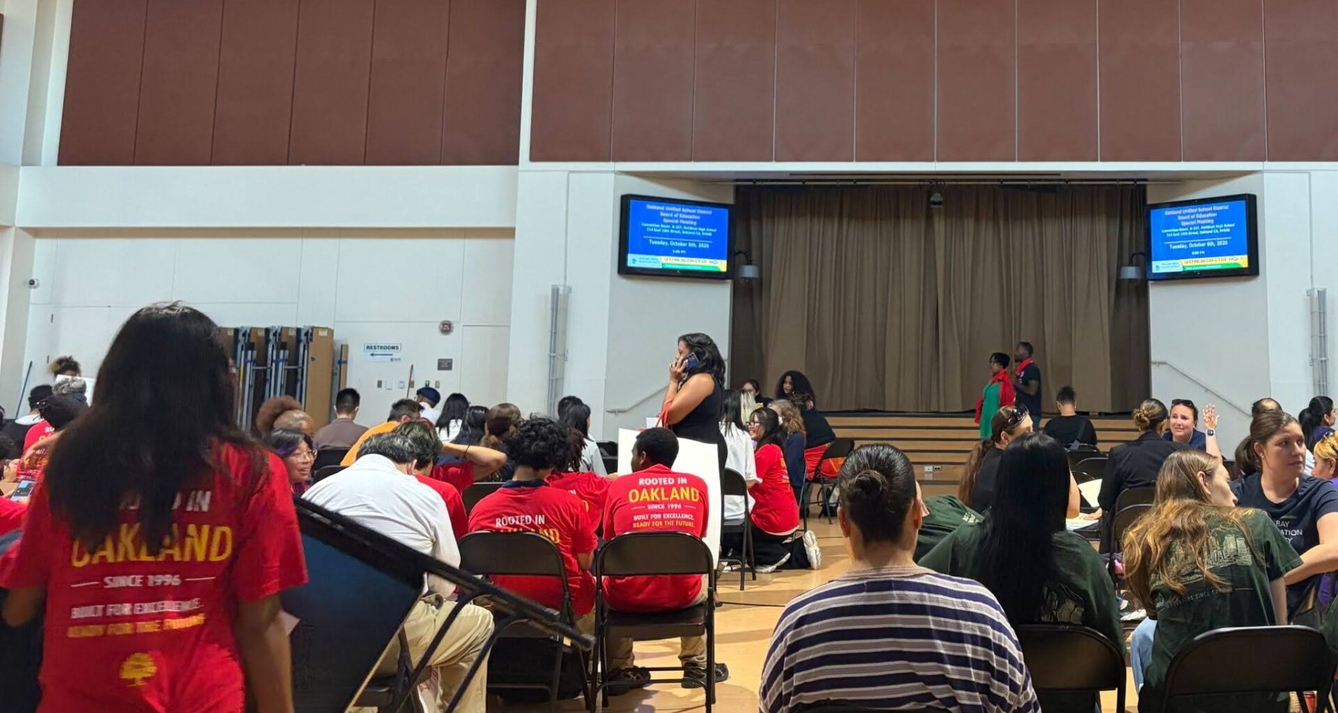 In a school gum about 50 or more people, many wearing the same red T-shirts, sit on folding chairs facing two digital screens.