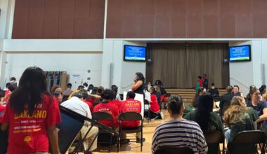 In a school gum about 50 or more people, many wearing the same red T-shirts, sit on folding chairs facing two digital screens.
