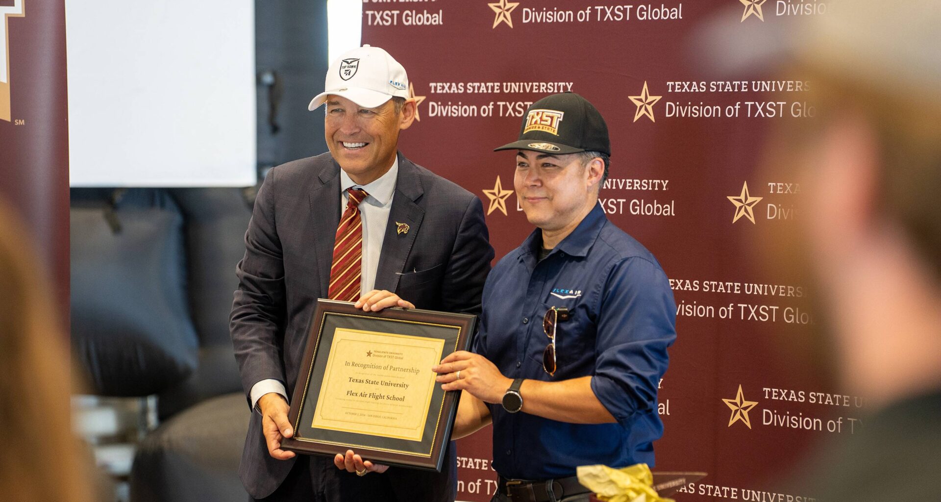 two men in baseball hats holding up a certificate in front of a texas state university step-and-repeat