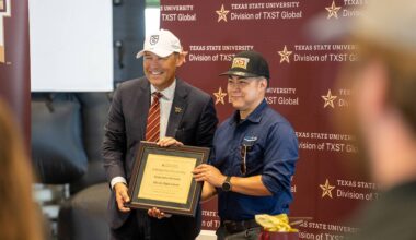 two men in baseball hats holding up a certificate in front of a texas state university step-and-repeat