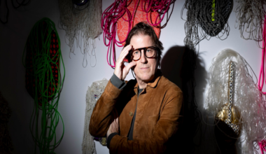 A man wearing glasses and a brown jacket sits on a bench, illuminated by a spotlight, with abstract artworks hanging on the wall behind him.