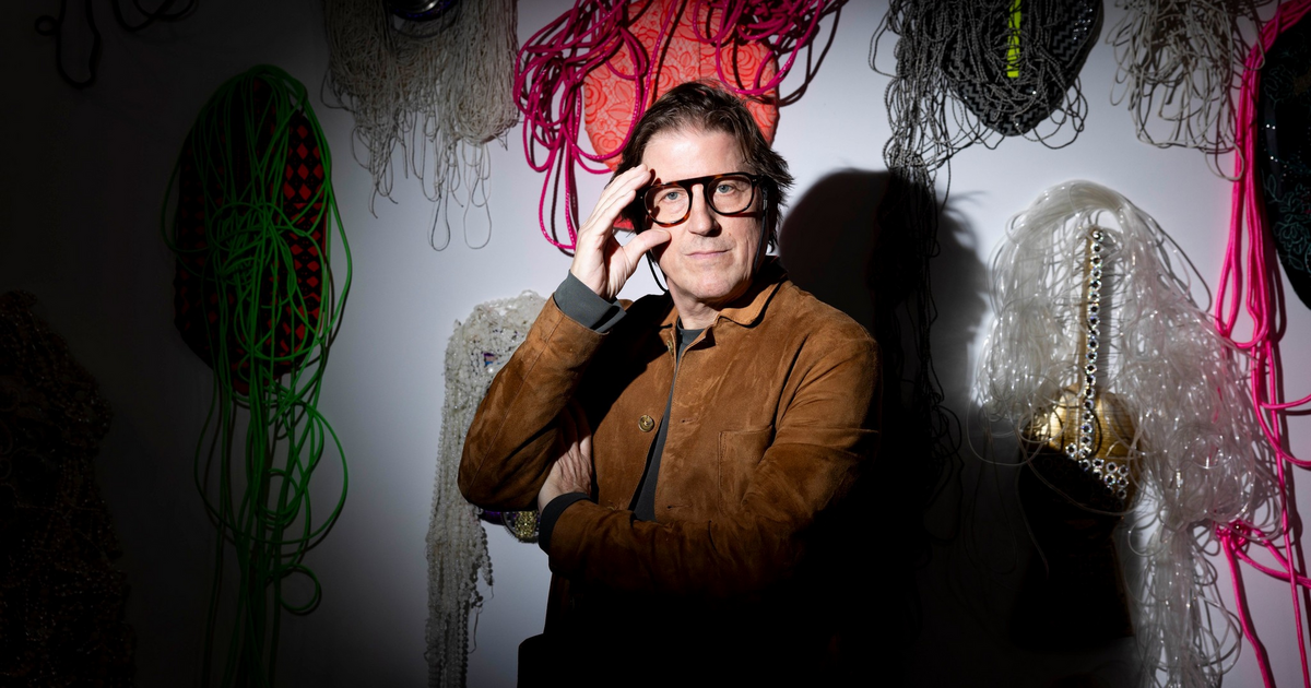 A man wearing glasses and a brown jacket sits on a bench, illuminated by a spotlight, with abstract artworks hanging on the wall behind him.