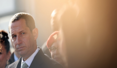 A man in a suit and tie looks thoughtfully to the side, surrounded by a blurred crowd with soft sunlight creating a warm glow.