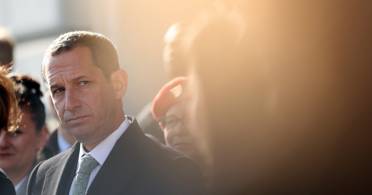 A man in a suit and tie looks thoughtfully to the side, surrounded by a blurred crowd with soft sunlight creating a warm glow.