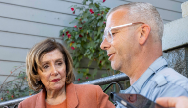 A woman in an orange blazer looks attentively at a man with gray hair and glasses speaking, while another hand holds a phone near them.
