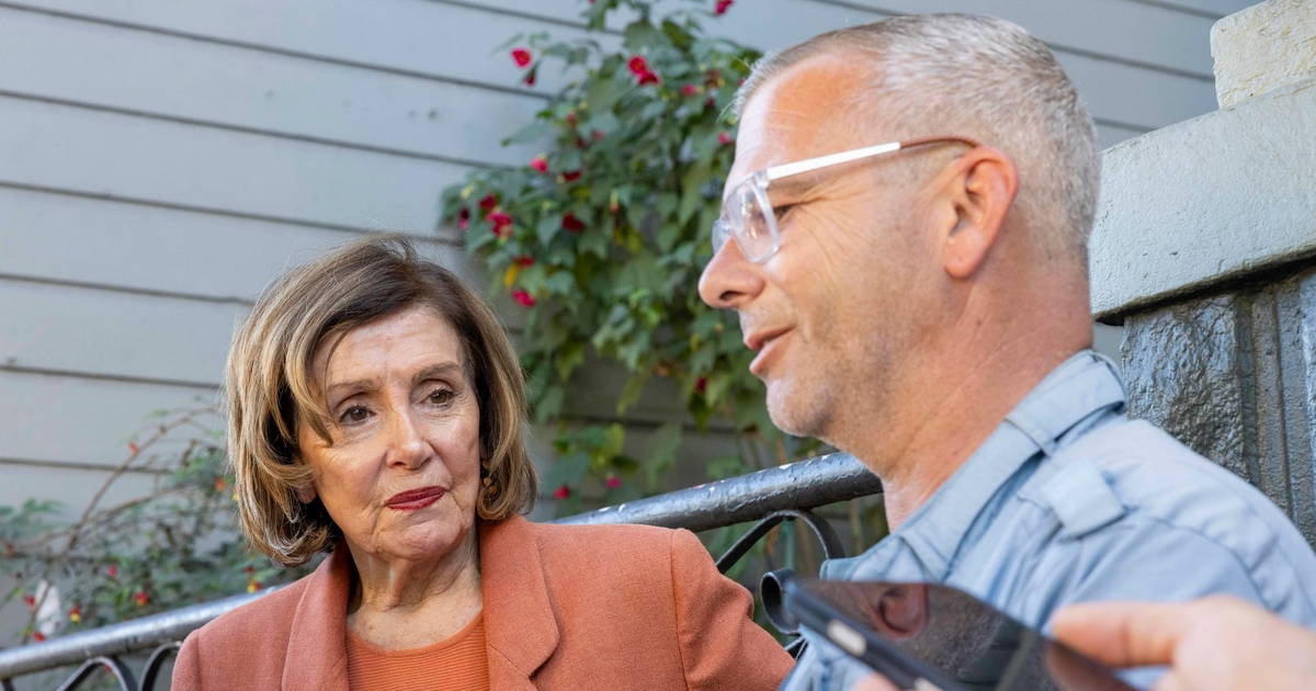 A woman in an orange blazer looks attentively at a man with gray hair and glasses speaking, while another hand holds a phone near them.