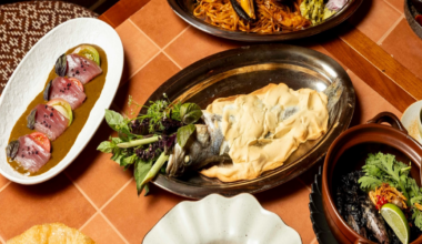 A table with assorted dishes including salt-crusted fish, seafood pasta, sliced tuna, rice, black stew, and a large crisp bread.