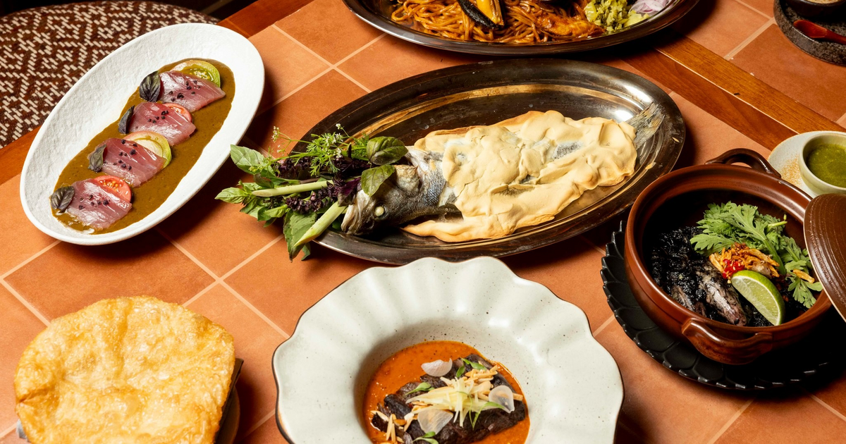 A table with assorted dishes including salt-crusted fish, seafood pasta, sliced tuna, rice, black stew, and a large crisp bread.