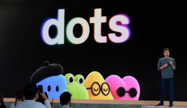 A presenter stands beside a large screen displaying the word "dots" in colorful letters and five colorful cartoon dots with various accessories and expressions.