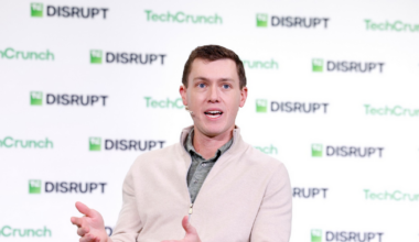 A man in a beige zip-up sweater and khaki pants gestures with his hands while speaking at a TechCrunch Disrupt event.