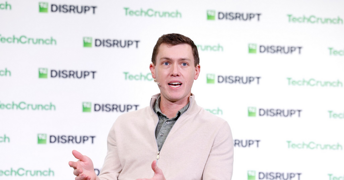 A man in a beige zip-up sweater and khaki pants gestures with his hands while speaking at a TechCrunch Disrupt event.