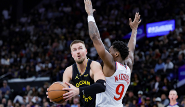 A Golden State Warriors player holds the basketball, preparing to shoot while a Philadelphia 76ers player with arms raised defends him.