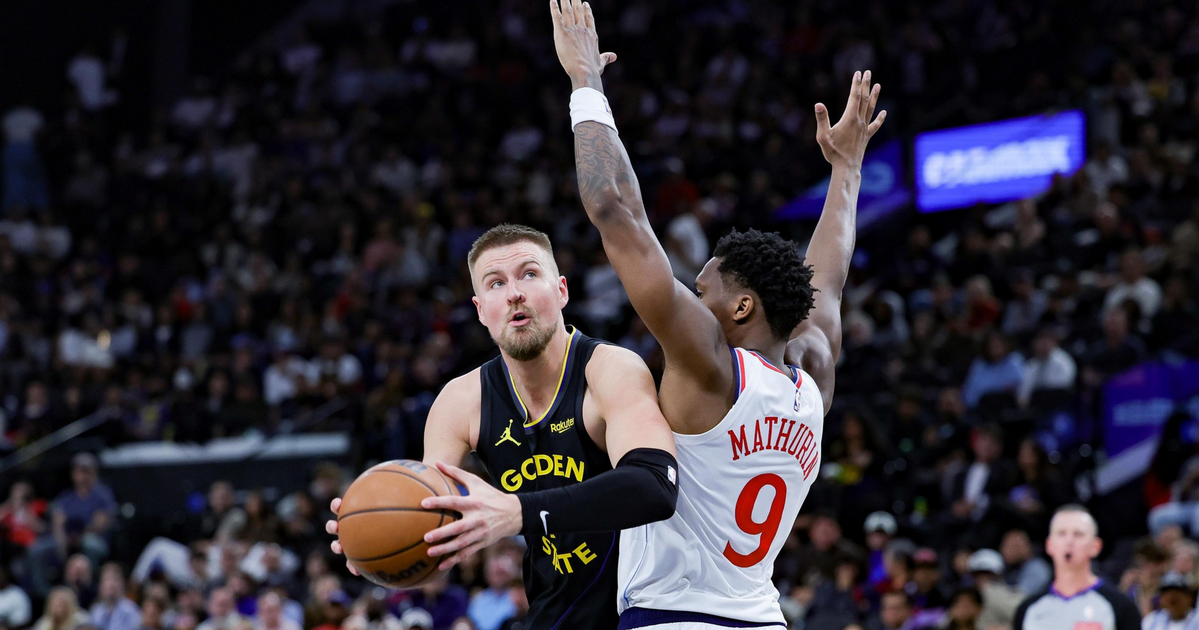 A Golden State Warriors player holds the basketball, preparing to shoot while a Philadelphia 76ers player with arms raised defends him.